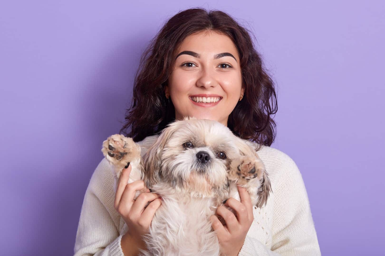 A smiling person is affectionately holding a small fluffy dog against a simple purple background. They both appear happy and content.