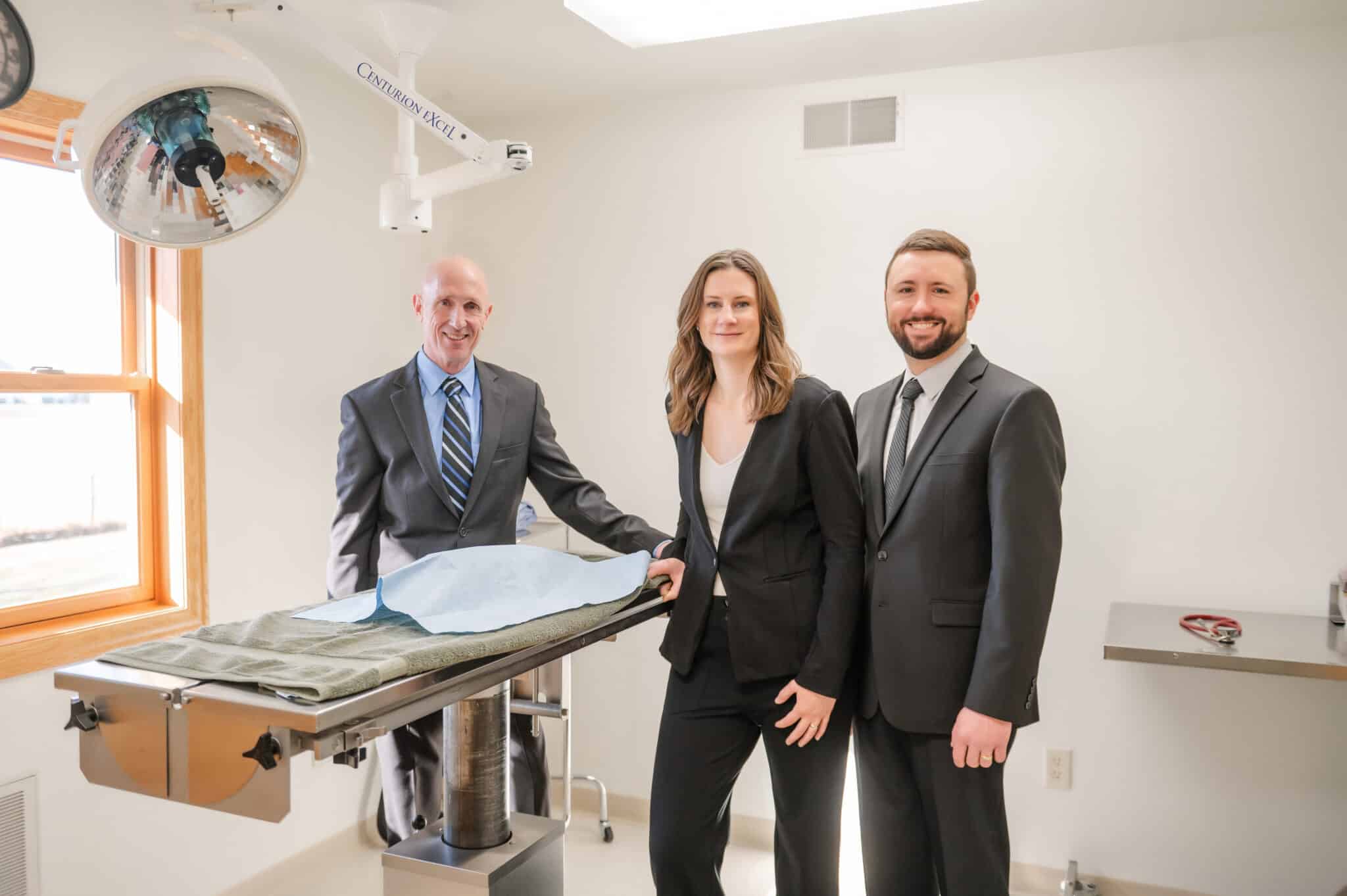 Three people in professional attire stand in a clinical room with an examination table, bright lighting, and a window on the left.