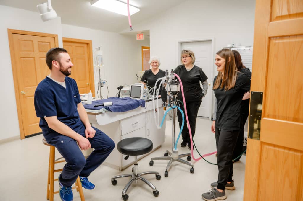 Four healthcare workers in scrubs are smiling and interacting in a bright, clean medical room with equipment like a monitor and IV stand.