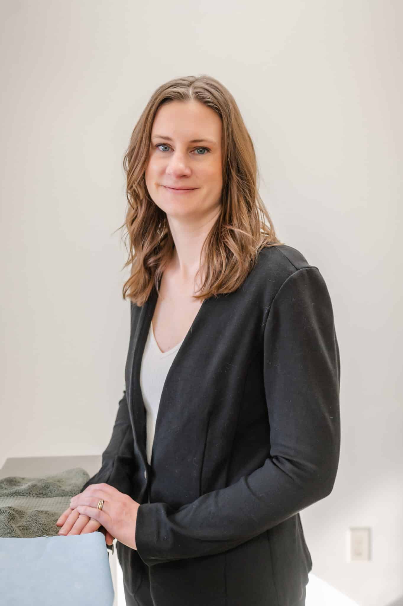 A person with shoulder-length hair, wearing a black blazer and a white top, is standing and smiling against a pale background.