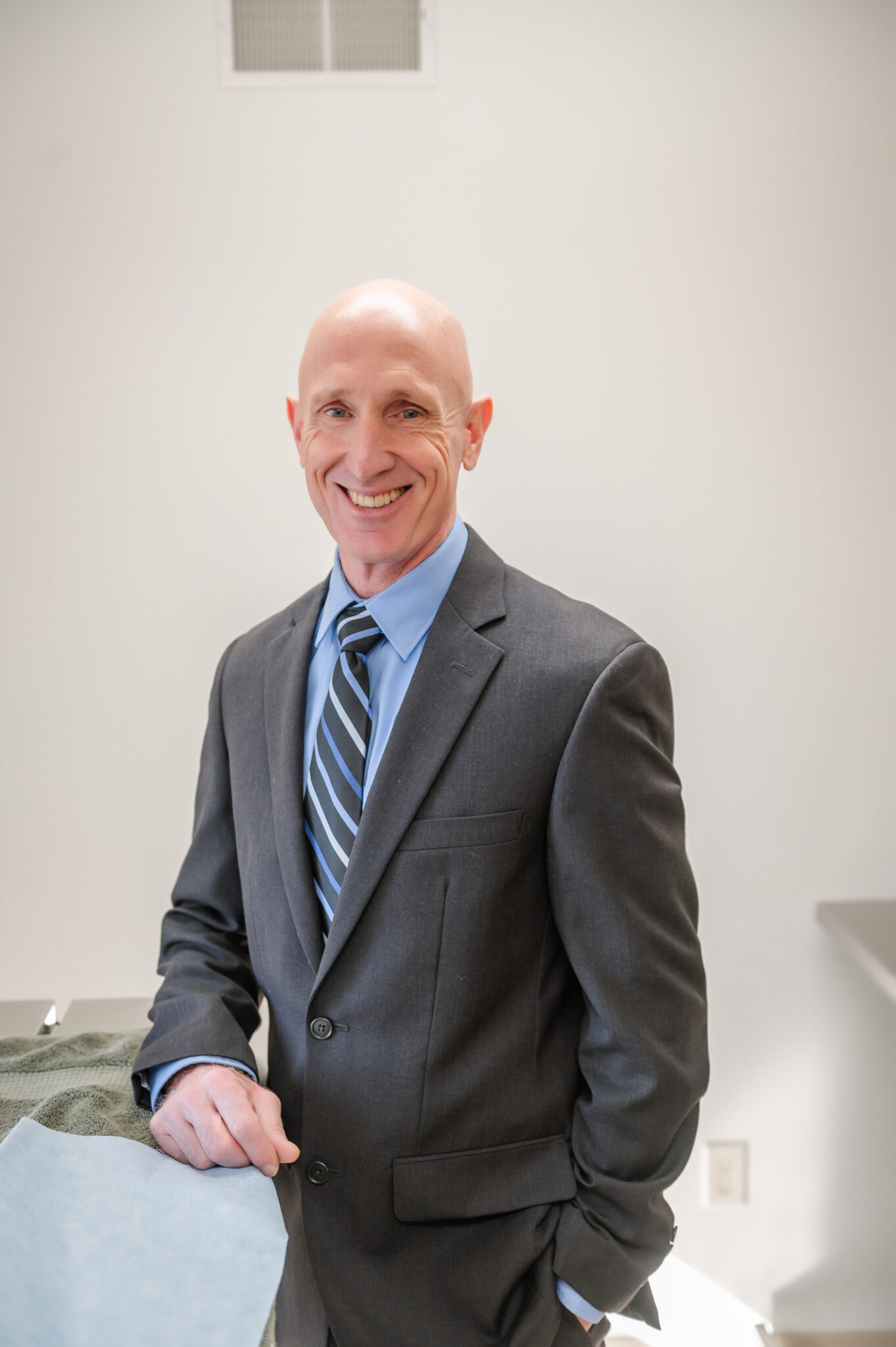 A smiling person in a dark suit with a blue tie stands in a room with a white background, leaning slightly against a table.