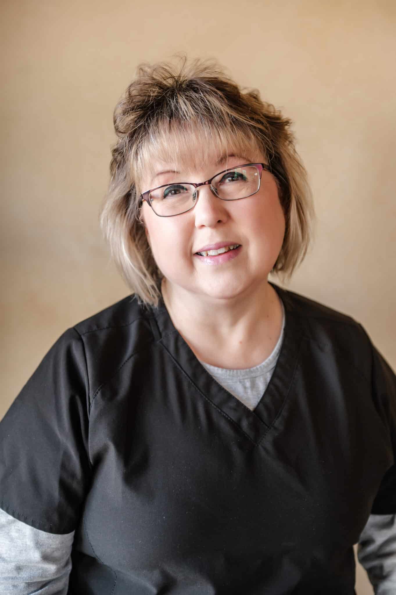 A smiling person with glasses, short hair, and a black shirt poses against a beige background. They appear professional, approachable, and friendly.