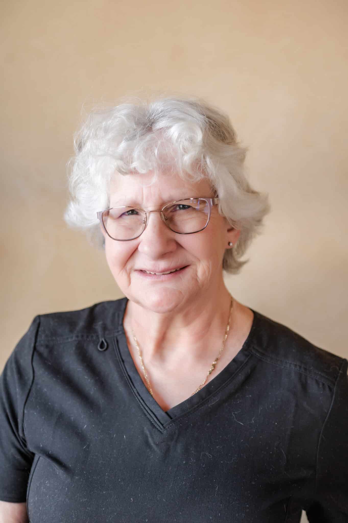 A smiling elderly person with curly white hair, glasses, and a black top stands against a beige background, exuding a warm and friendly demeanor.